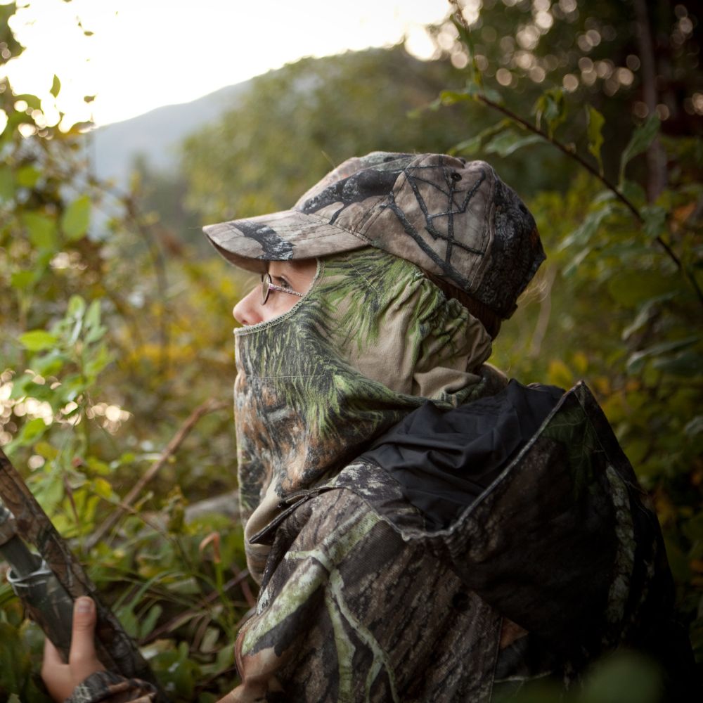 Person in camouflage clothing and hat holding a rifle in a forest setting