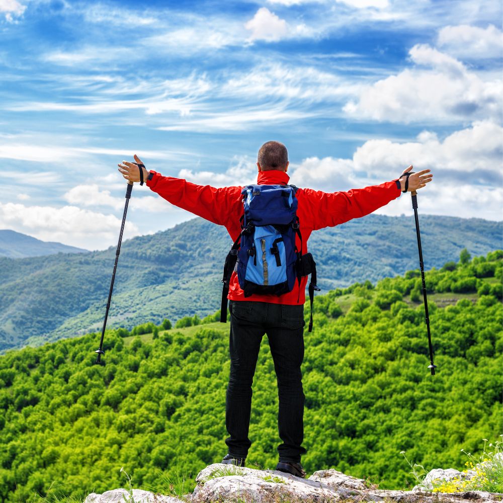 Hiker in a red jacket with a blue backpack standing on a mountain top with arms outstretched.