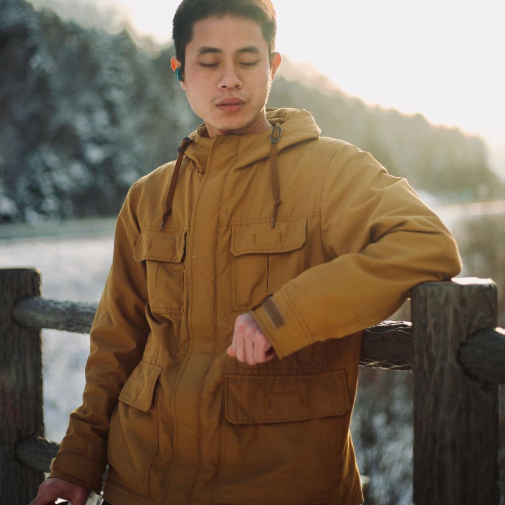 Man wearing a brown jacket standing by a wooden railing with a scenic background