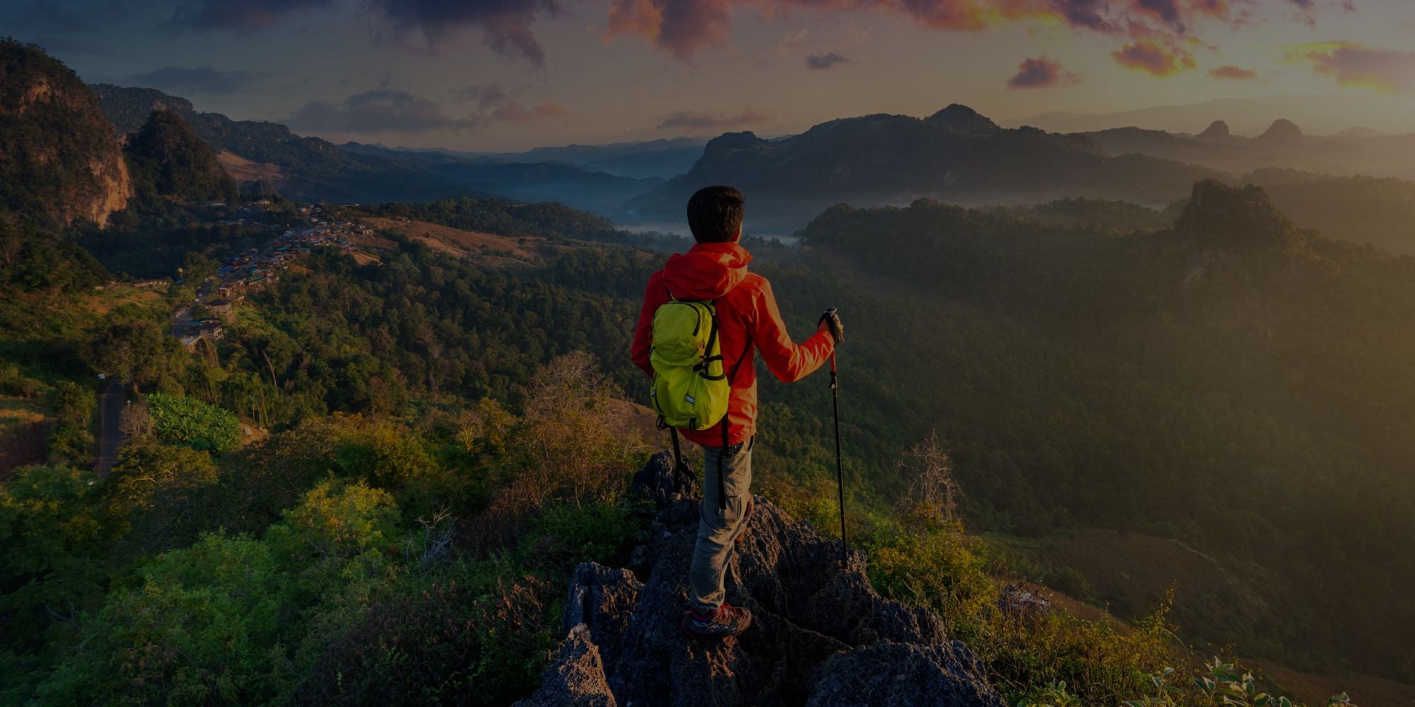 Person with a backpack and hiking pole standing on a mountain peak overlooking a scenic landscape.