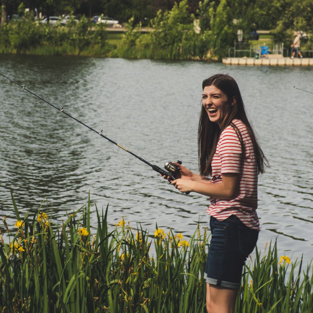 Woman fishing by a lake with greenery and water in the background