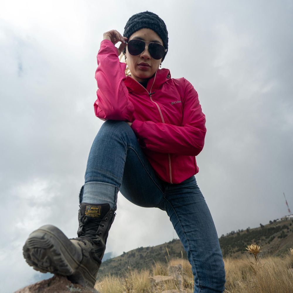 Person in a pink jacket and sunglasses sitting on a rock with a mountainous background